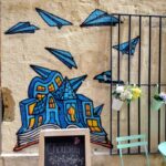 Streetart in Montpellier: Ein Haus steht auf einem aufgeschlagenen Buch. Um es herum schwirren Papierflieger.